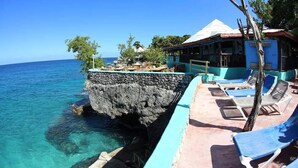 Un hotel frente al mar con muro de piedra, aguas azul turquesa y sillas de descanso.