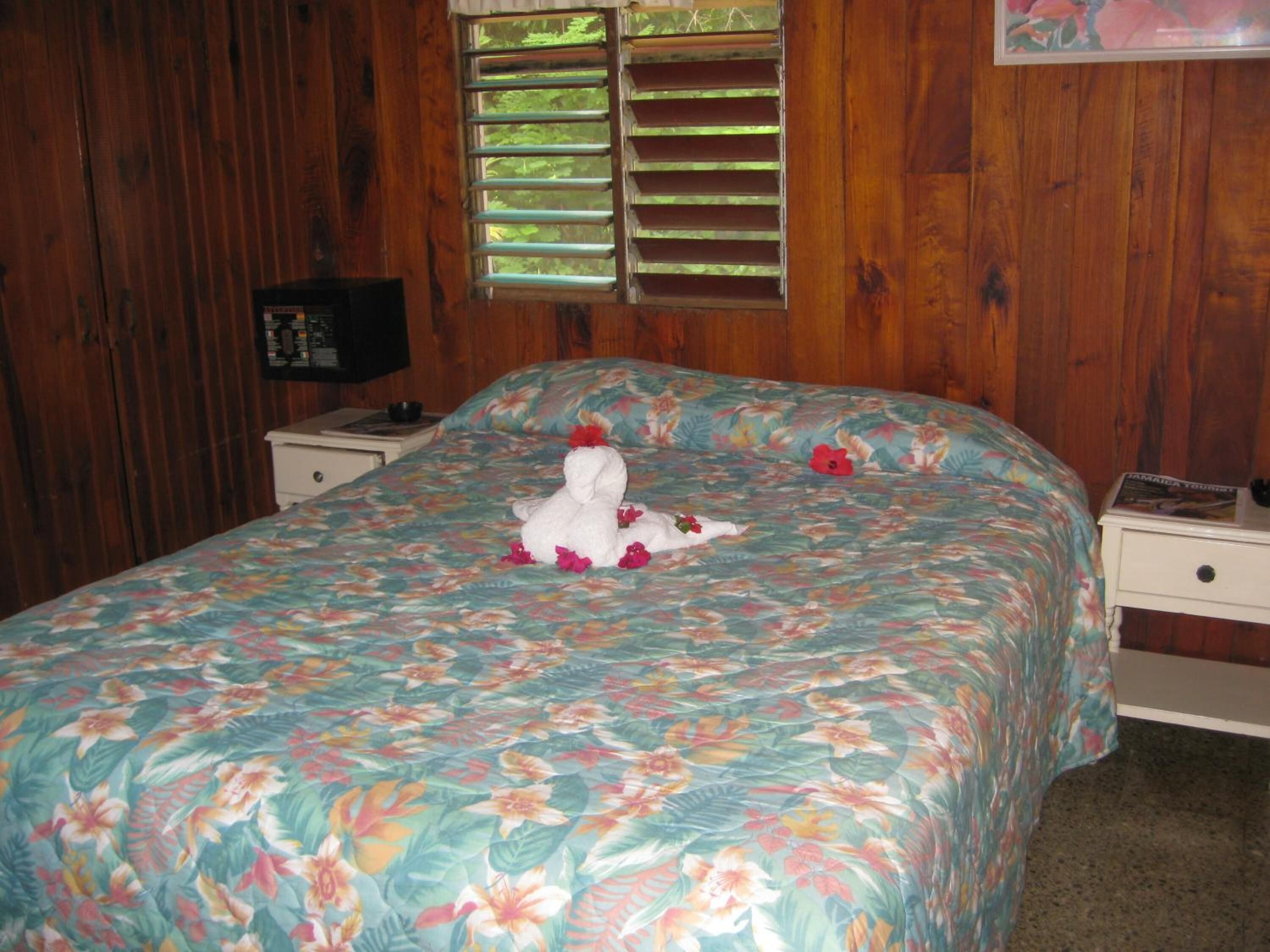 A bedroom with a floral bedspread, a white stuffed animal on the bed, and a window with blinds.