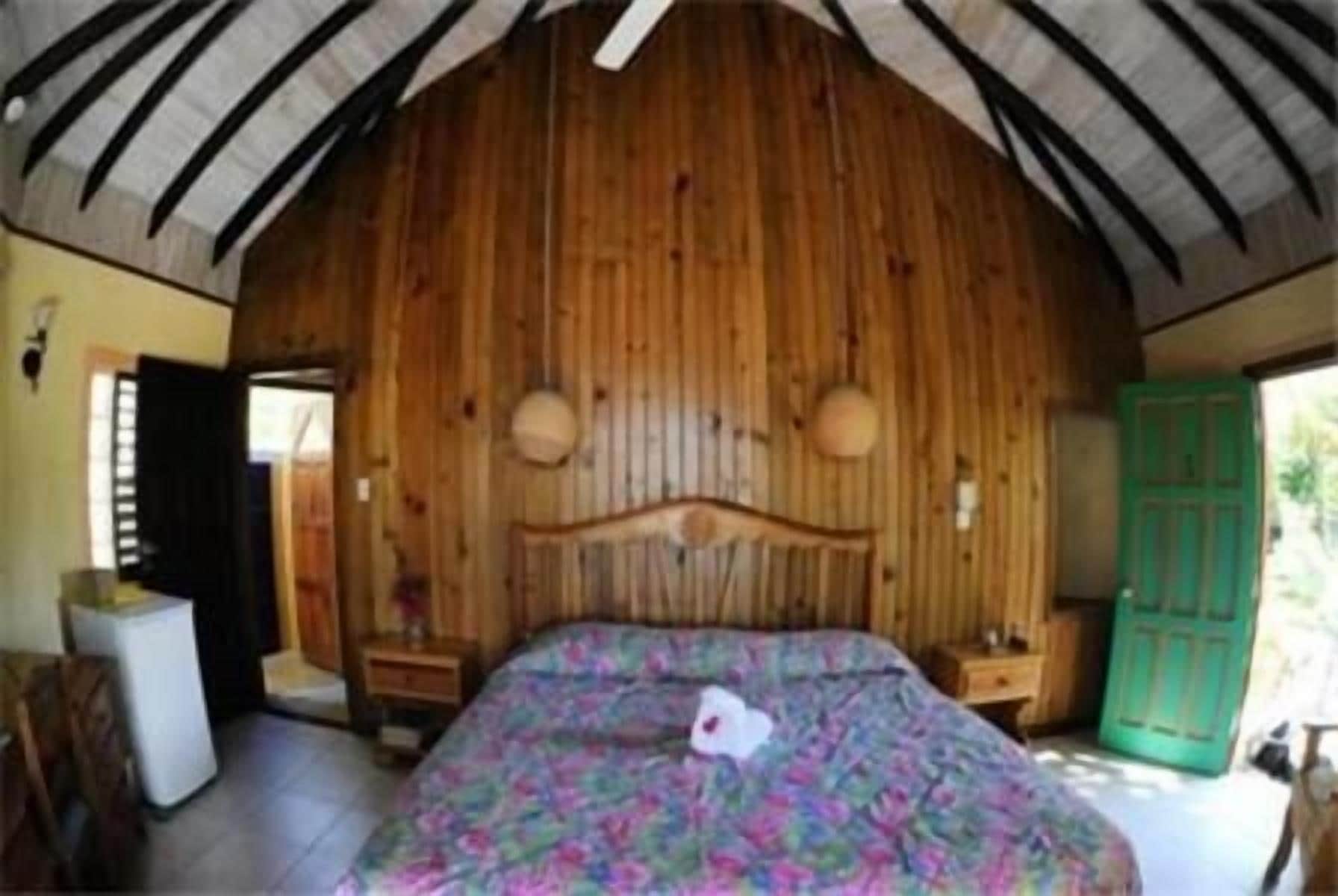 A bedroom with a wooden ceiling, a bed with a floral bedspread, a nightstand, and a green door.