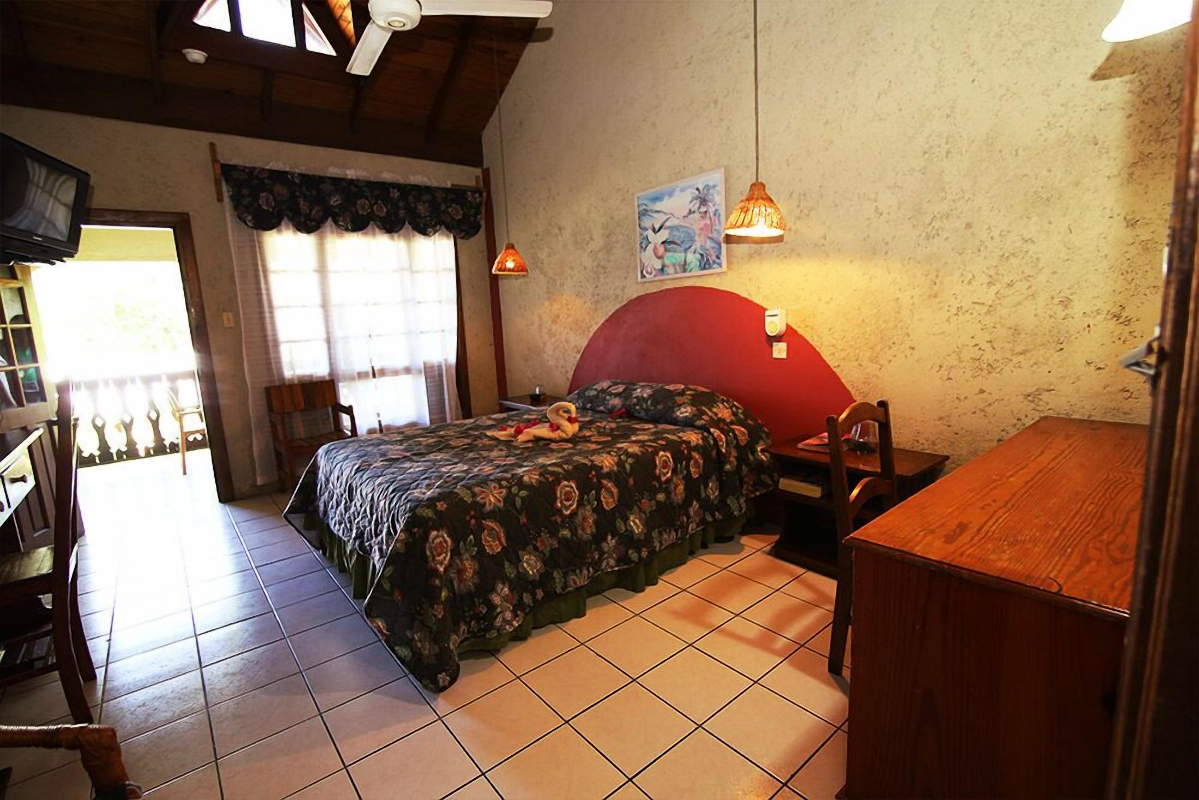 A bedroom with a bed, a desk, a chair, a wooden table, and a window with curtains.