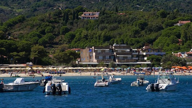 A coastal area with boats docked near a beach, a hotel building, and a hillside with trees. - Hotel Select (Campo nell'Elba)