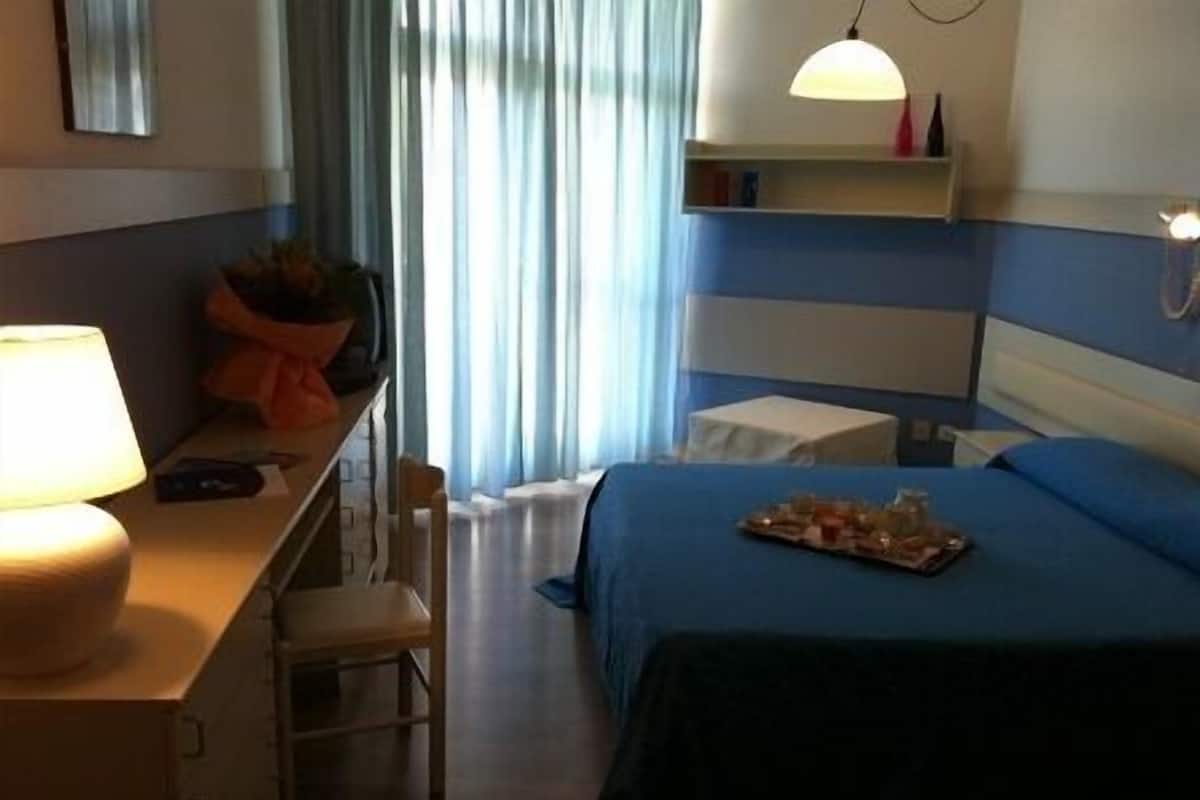 A hotel room with a bed, a desk with a lamp, a chair, a bedside table, a window with curtains, and a shelf with a lamp and decorative items.