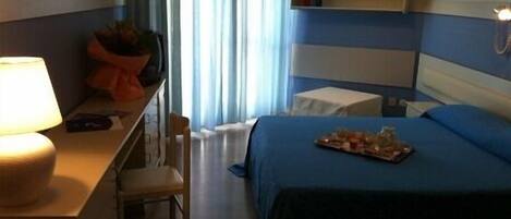 Habitación de hotel con cama, escritorio con lámpara, silla, mesita de noche, ventana con cortinas y estante con lámpara y objetos decorativos.