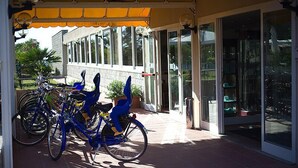 A covered outdoor area with bicycles parked under a yellow awning, a potted plant, and a building with large windows and a glass door entrance. - Hotel Select (Campo nell'Elba)