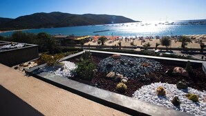 A landscaped garden with a variety of plants and rocks, a beach with umbrellas and people, and a view of the sea and mountains in the background. - Hotel Select (Campo nell'Elba)