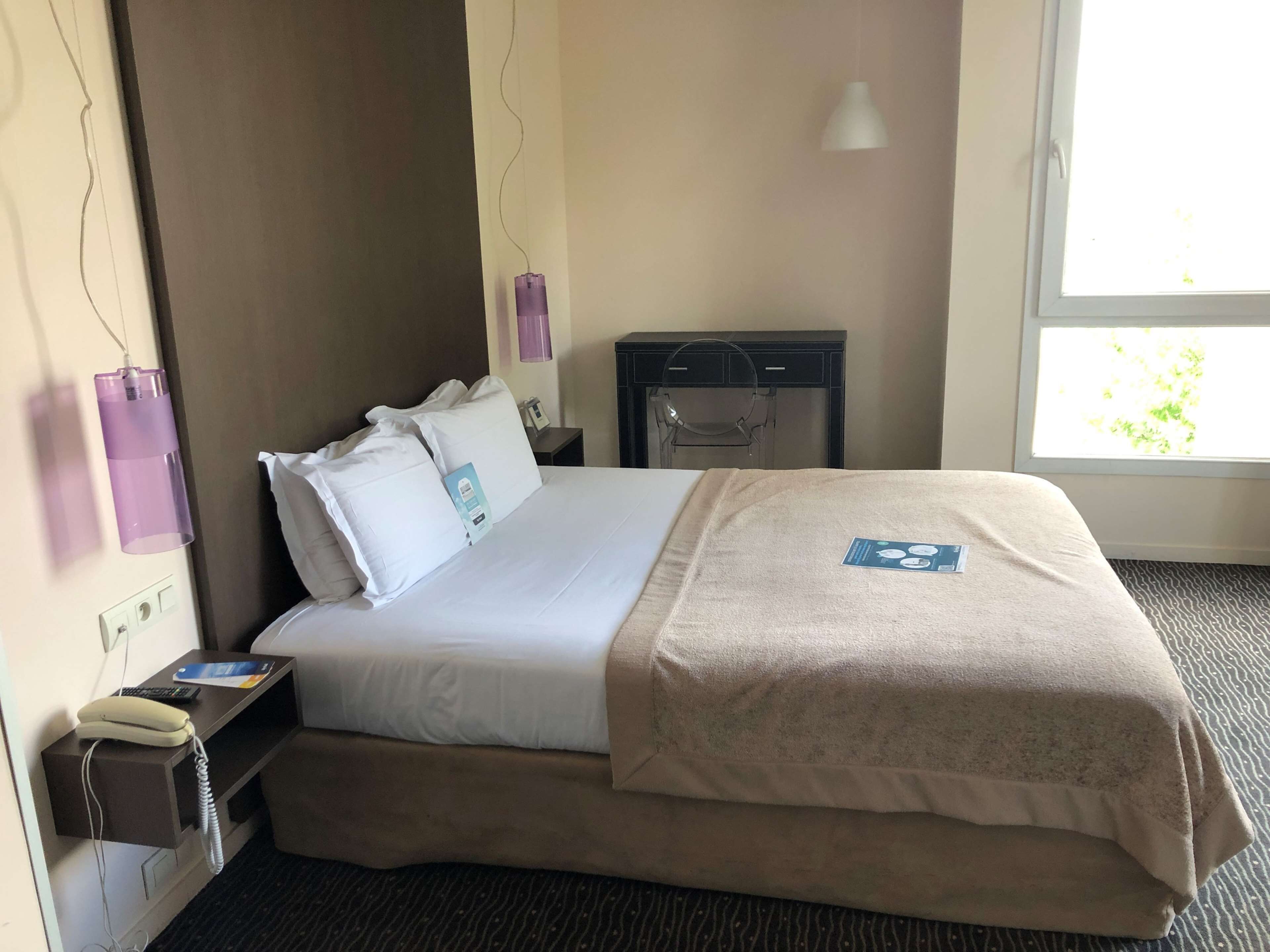 A hotel room with a bed, bedside table, lamp, and a window with a view of greenery.