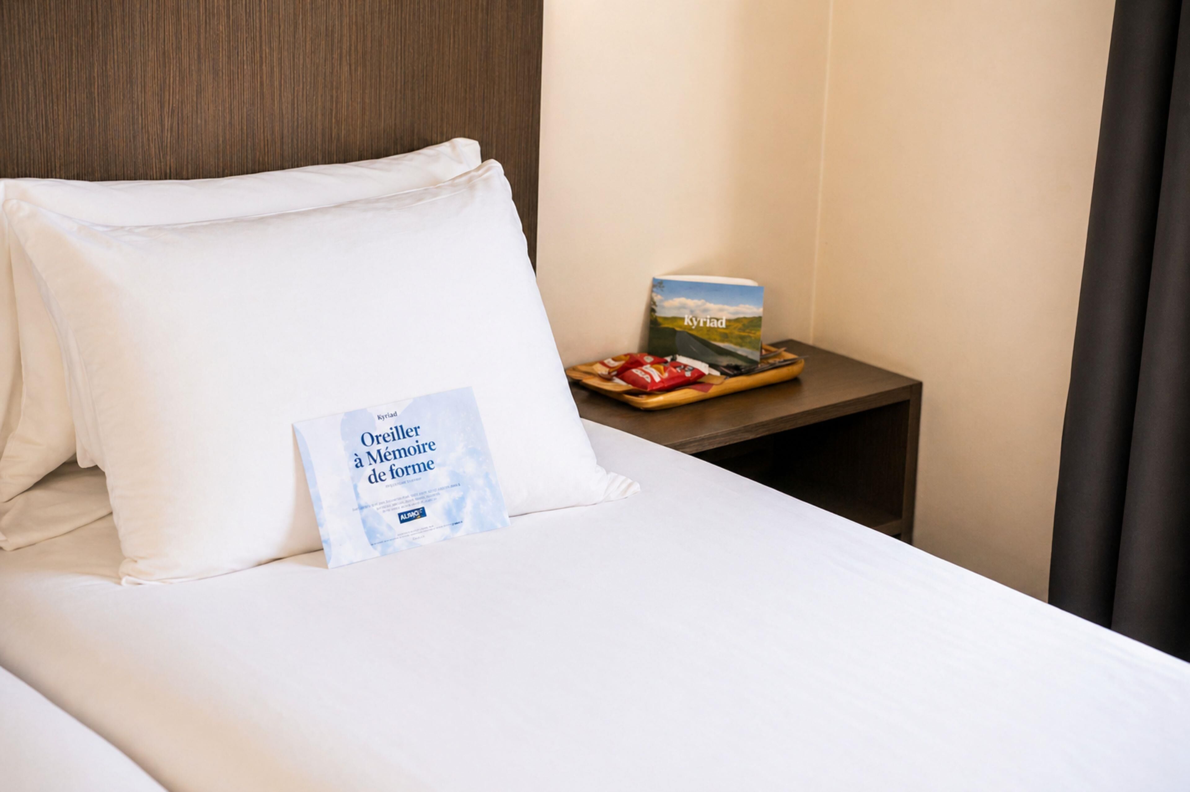 A neatly made bed with white linens, a pillow, a book titled 'Kyriad,' and a tray with snacks on a bedside table.