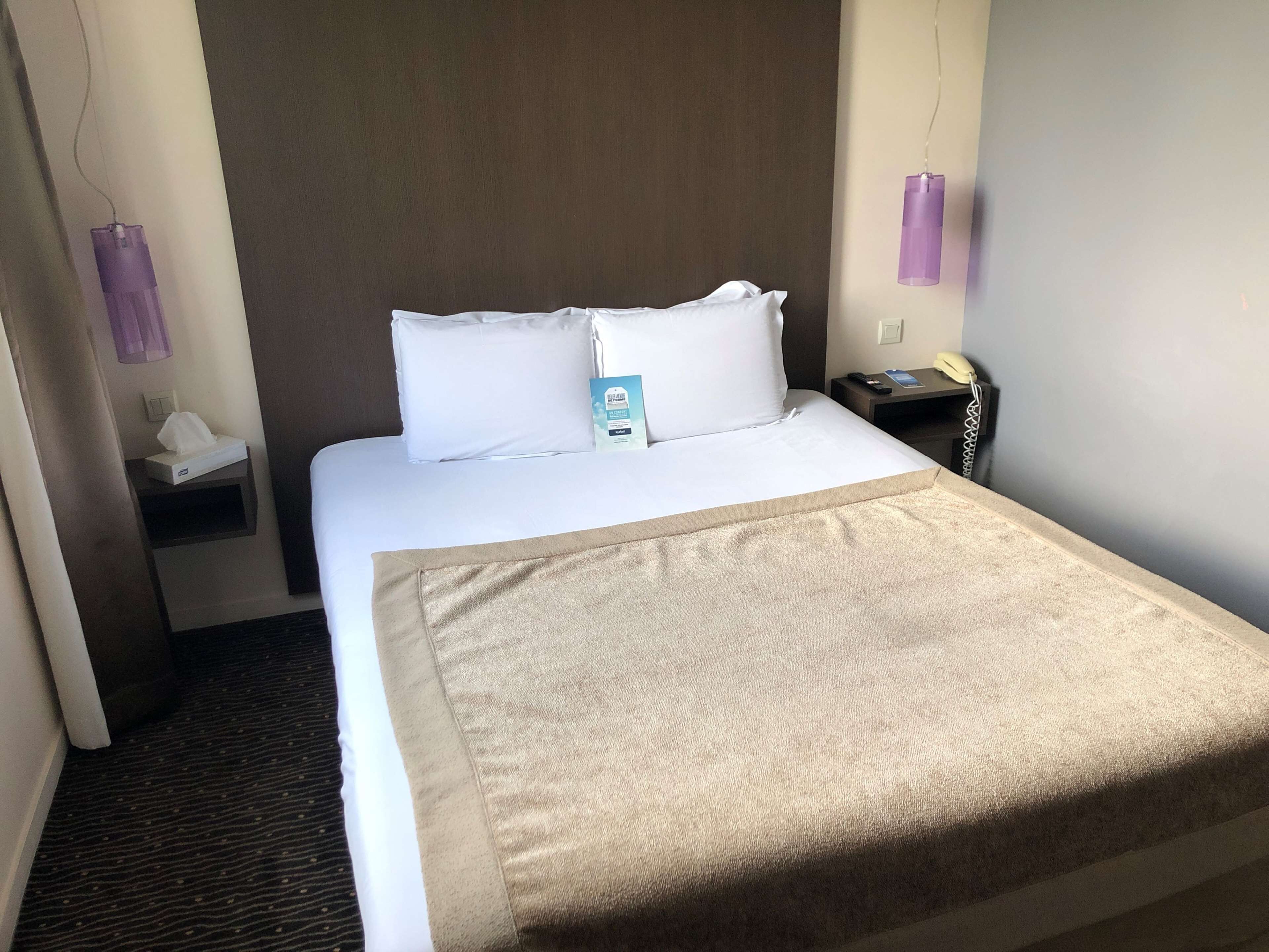 Habitación de hotel con una cama grande, almohadas blancas, un plaid color beige, una mesita de noche con una lámpara, un teléfono y una caja de pañuelos.