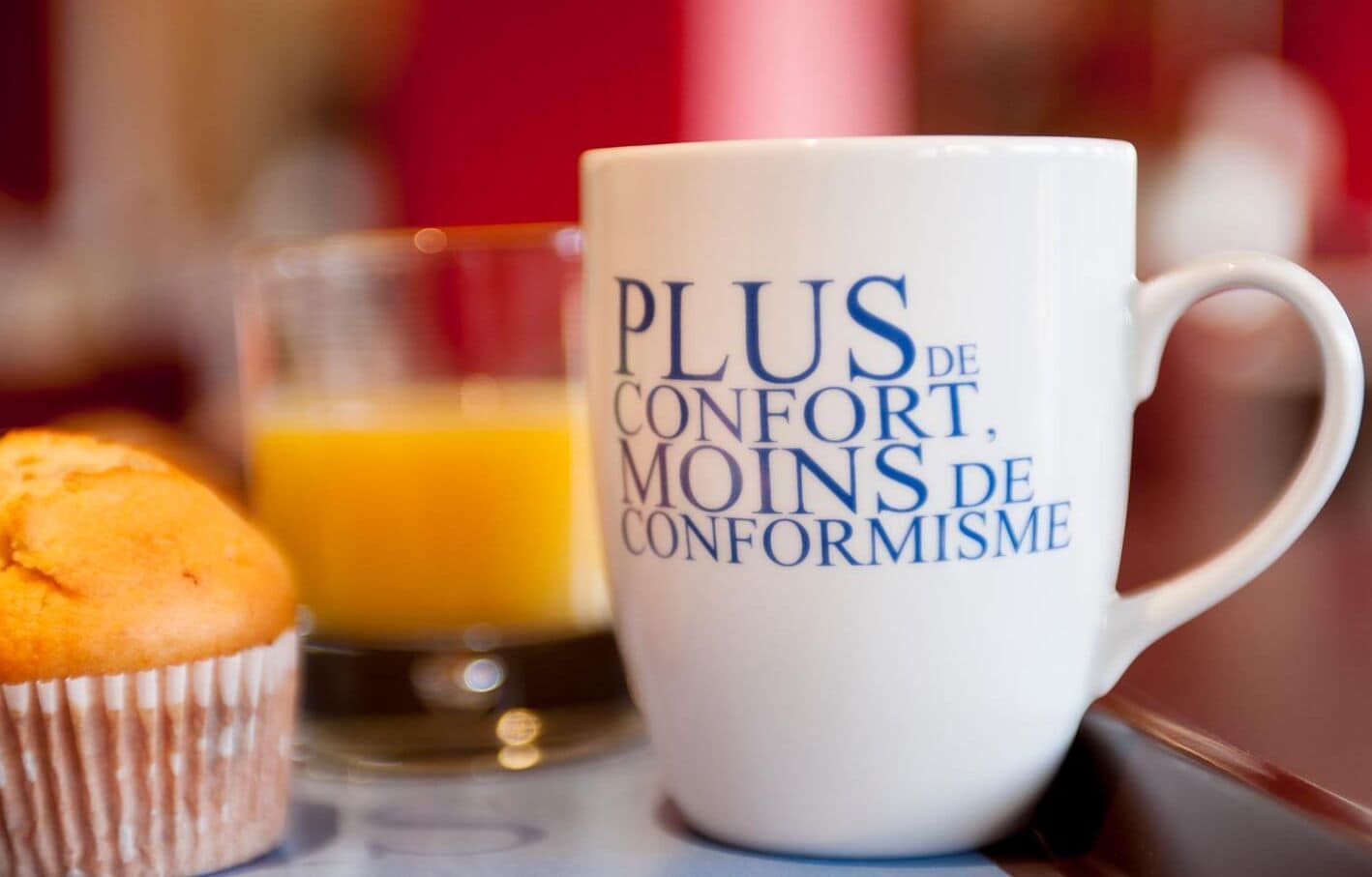 Eine weiße Tasse mit Schriftzug, ein Muffin und ein Glas Orangensaft auf einem Tisch.