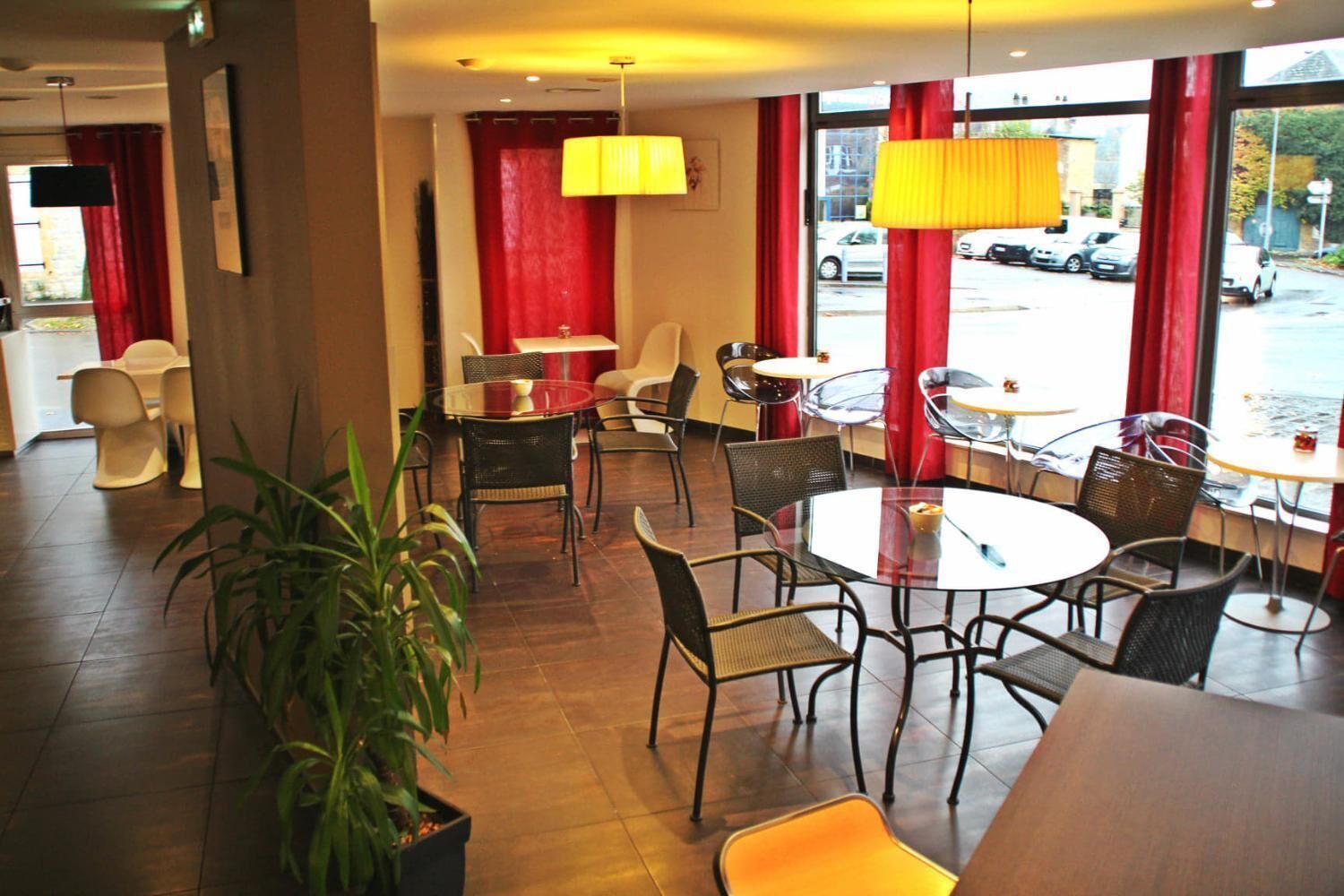 A modern restaurant with round tables and chairs, large windows with red curtains, and a view of the street outside.
