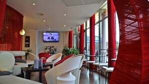 Ein moderner Lounge-Bereich mit weißen Sofas, roten Vorhängen und einem Fernseher, der eine Nachrichtensendung zeigt.