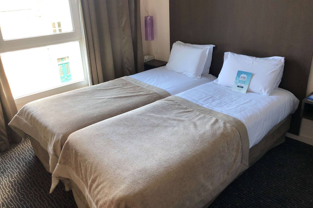 Habitación de hotel con dos camas, una ventana con cortinas, una mesita de noche y una lámpara fijada en la pared.