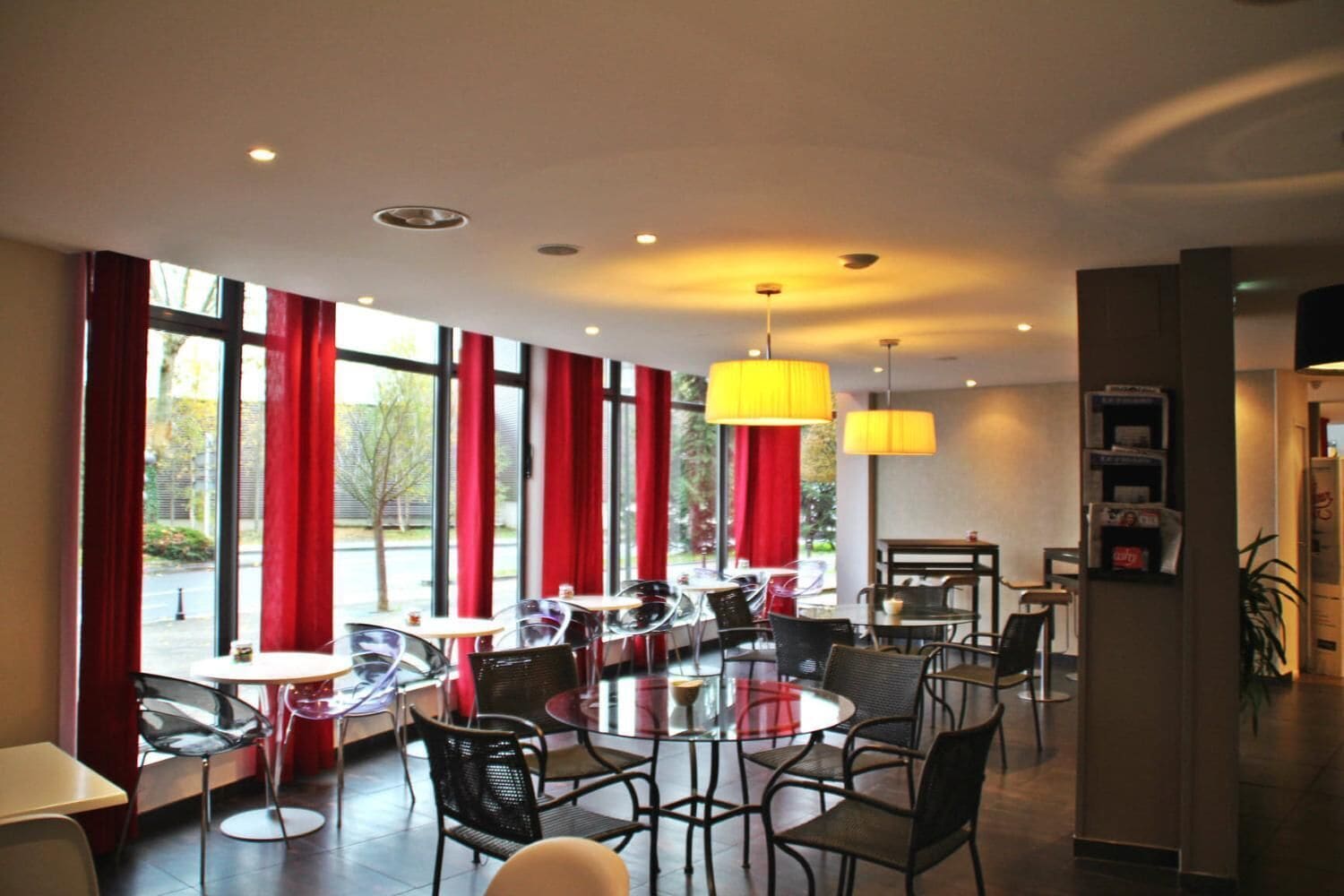 A modern dining area with round tables and chairs, large windows with red curtains, and yellow pendant lights.