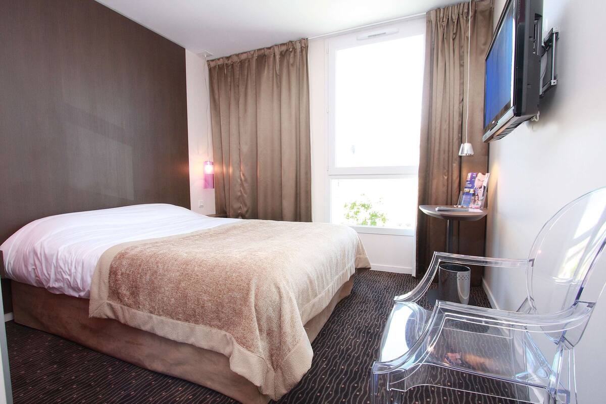 Una habitación de hotel con cama, televisor montado en la pared, una mesita de noche con lámpara y una silla.