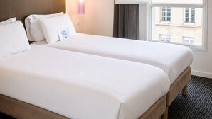 Ein Hotelzimmer mit zwei Betten, einem Fenster mit Vorhängen, einer Wandleuchte und Blick auf Gebäude.