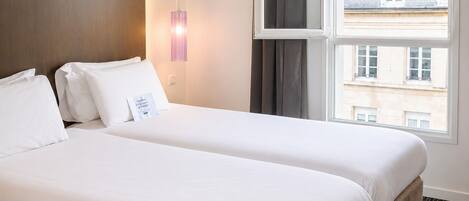 Habitación de hotel con dos camas, una ventana con cortinas, una lámpara de pared y vistas a edificios.