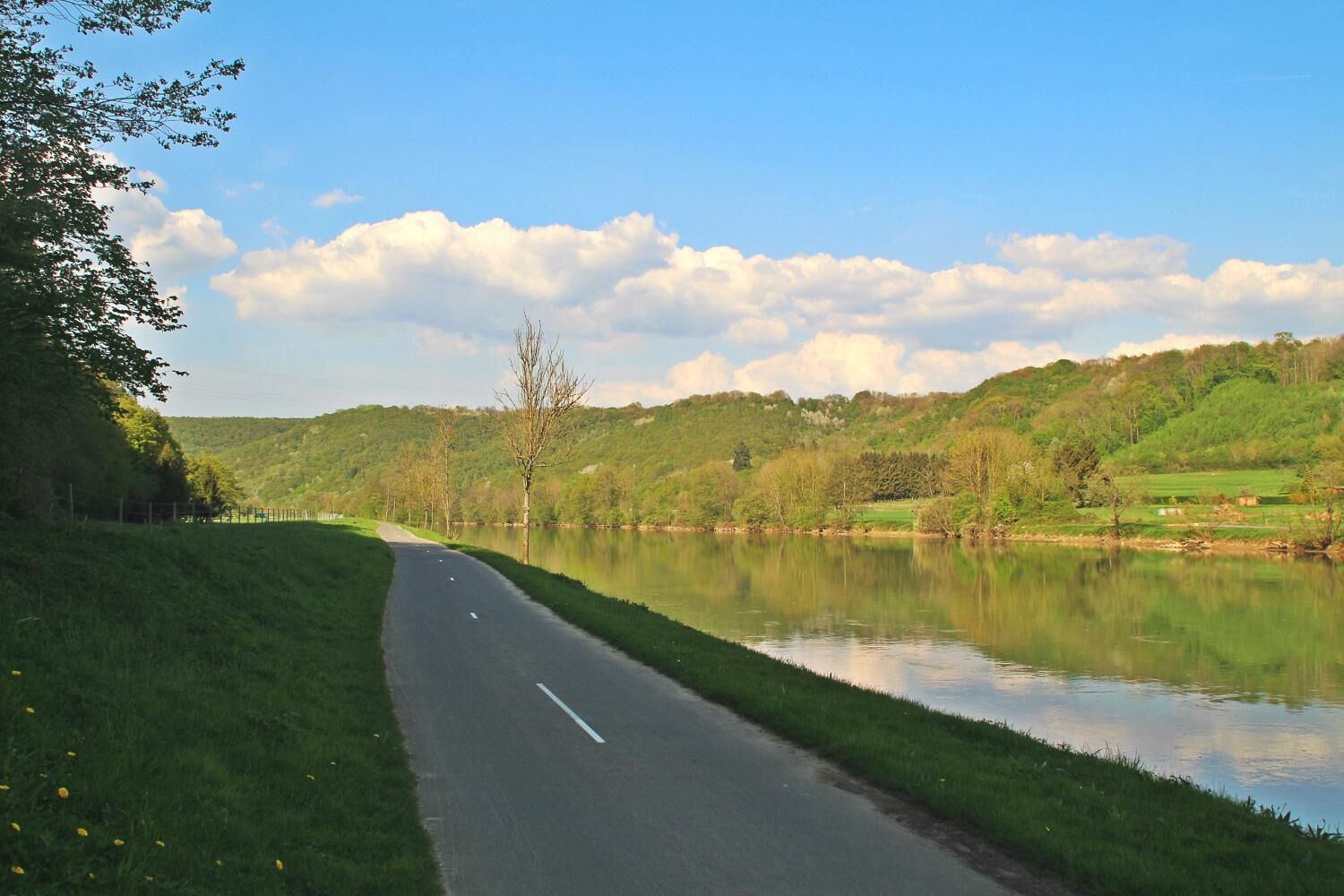 Una carretera serpentea junto a un lago tranquilo con orillas cubiertas de hierba y árboles, bajo un cielo parcialmente nublado.