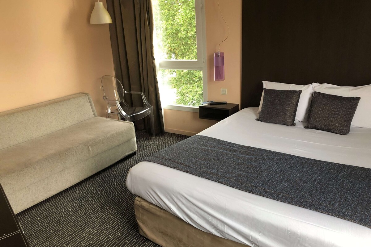 Une chambre d’hôtel comprenant un lit, un canapé, une chaise, une fenêtre avec des rideaux et une table de chevet avec une lampe.