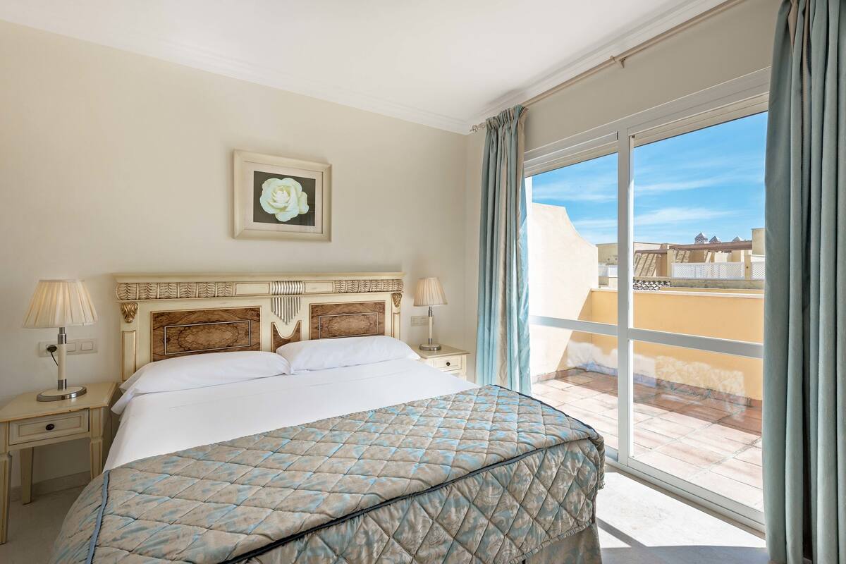 Ein Hotelzimmer mit einem großen Bett, einem Nachttisch, einer Lampe, einem gerahmten Bild an der Wand und Blick auf die Stadt durch das Fenster.