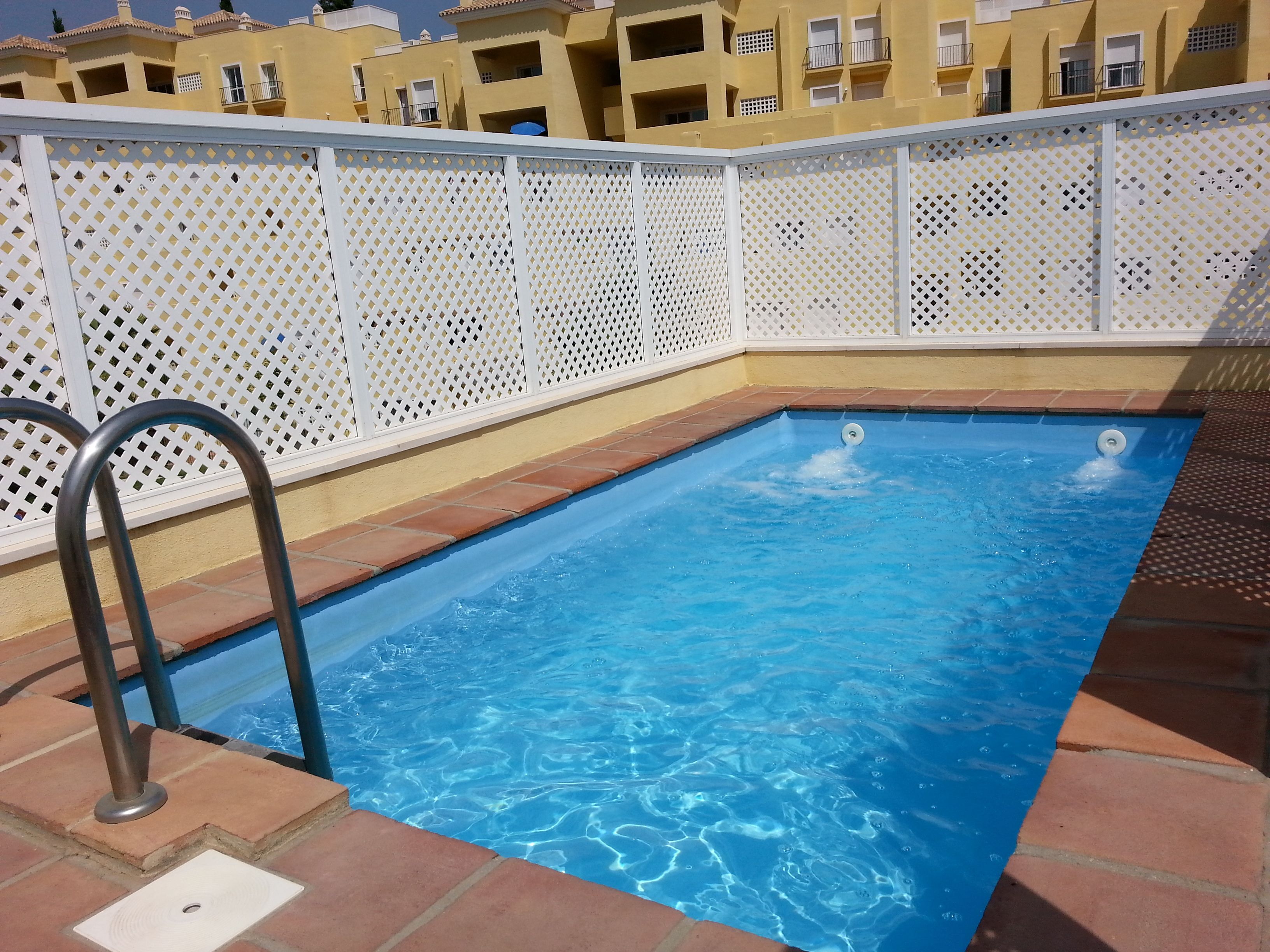 Et rektangulært swimmingpool med krystalklart, blå vand, omgivet af et hvidt gitrehegn og terracotta-fliser.