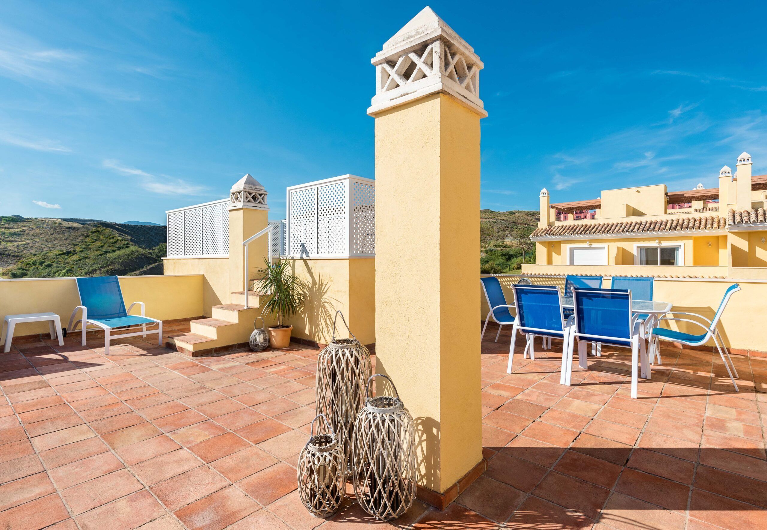 Una terraza con sillas azules, una mesa blanca y un jarrón decorativo.