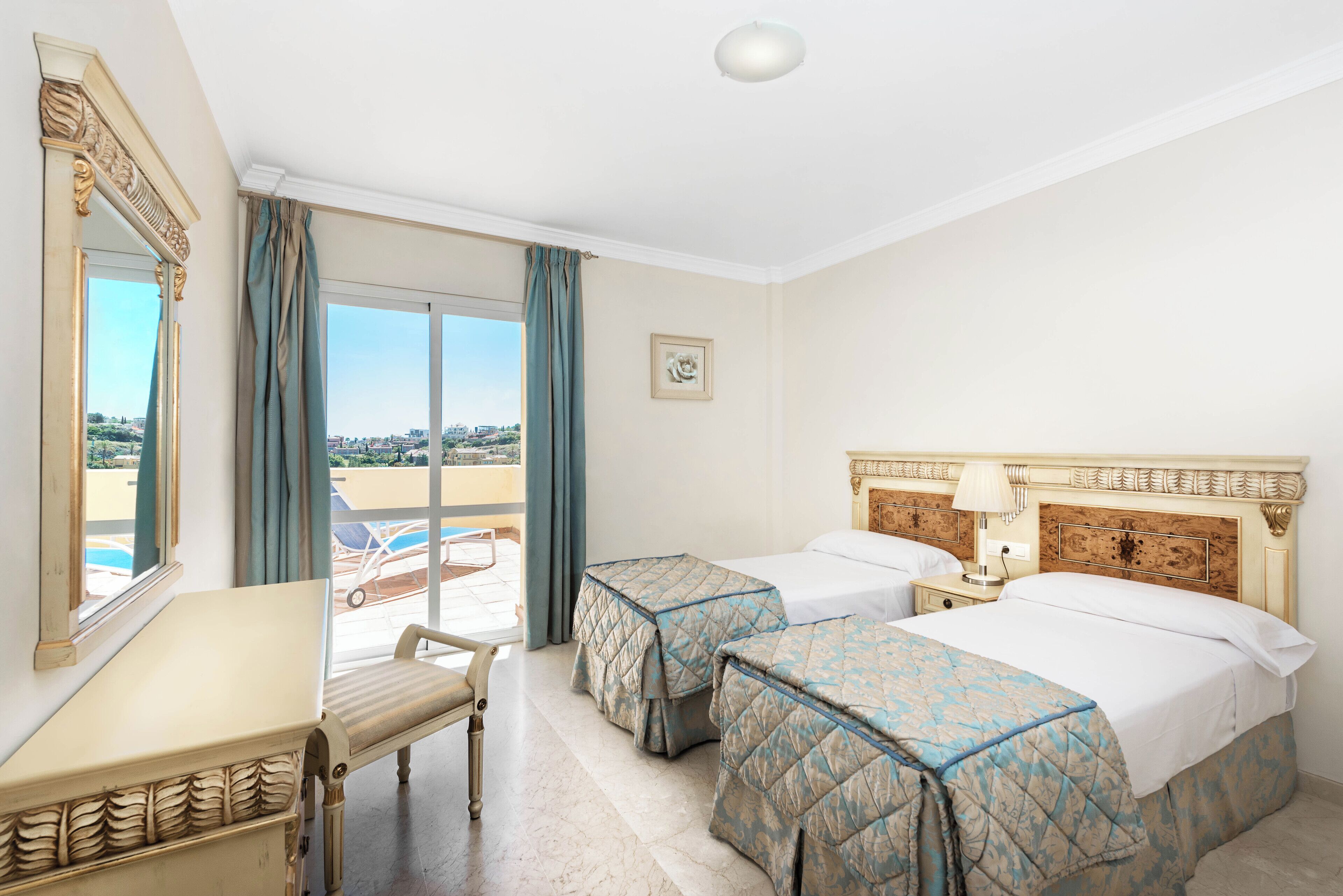 A hotel room with two beds, a desk, and a chair. There is a large window with curtains, a picture on the wall, and a view of the outside.