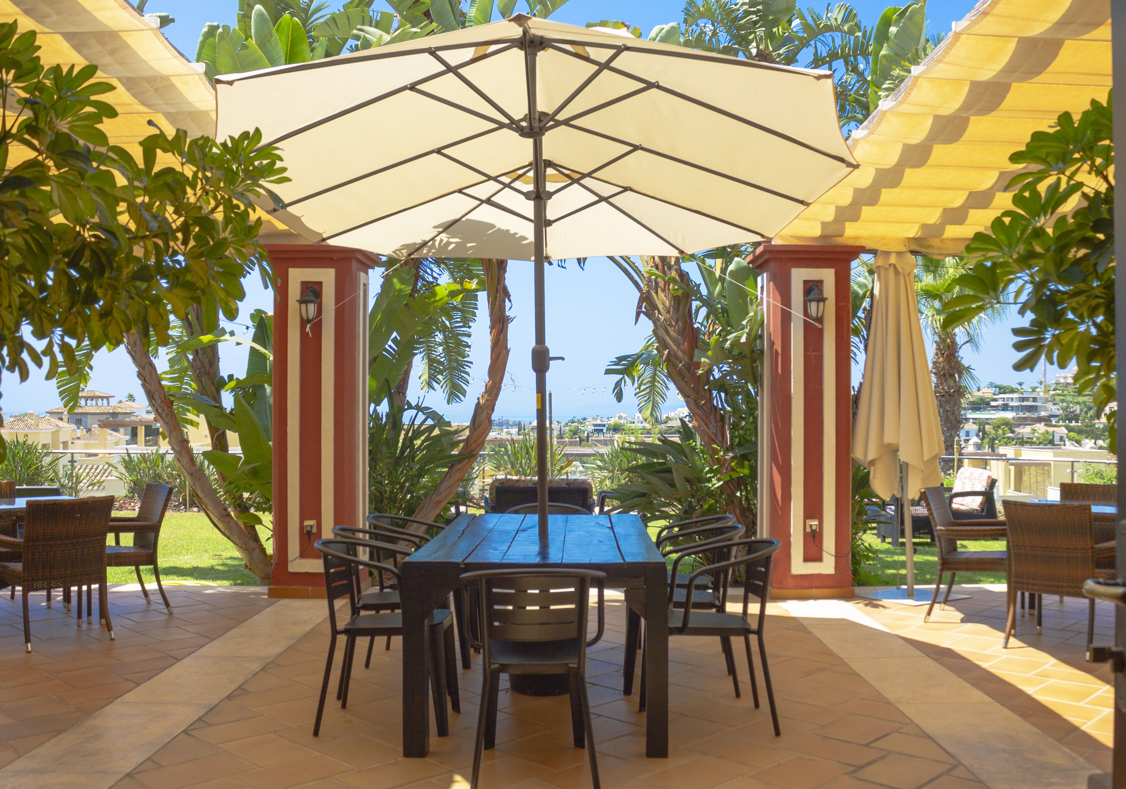 Eine Terrasse im Freien mit einem Holztisch und Stühlen, umgeben von Pflanzen und Sonnenschirmen.