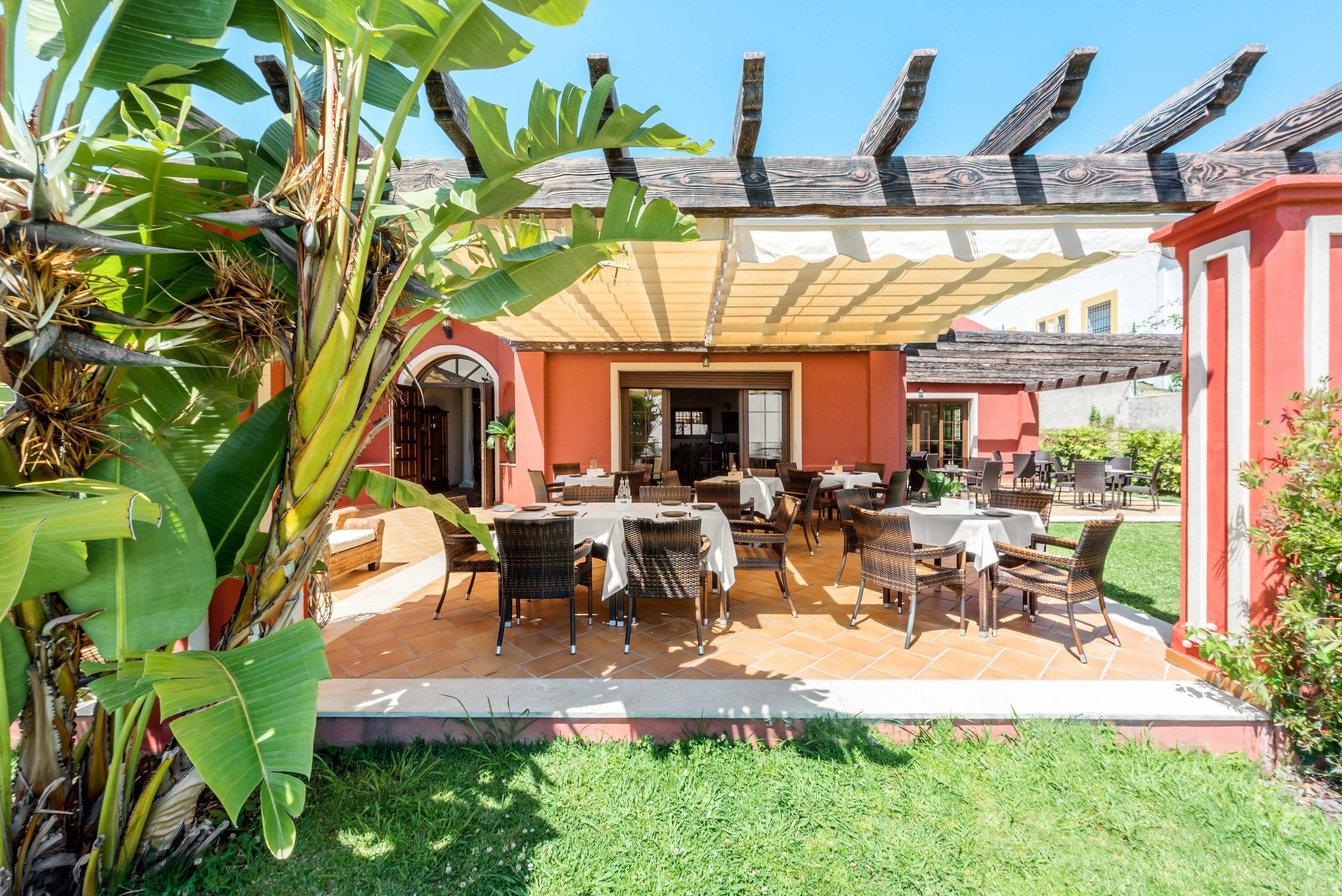 An outdoor patio with tables and chairs, surrounded by lush greenery and a building with a tiled roof.