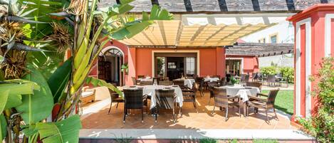 An outdoor patio with tables and chairs, surrounded by lush greenery and a building with a tiled roof.