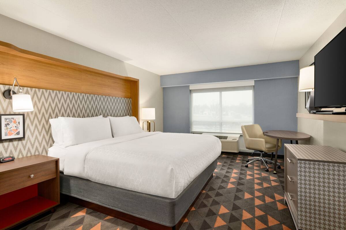 A hotel room with a bed, bedside table, lamp, TV, desk, chair, and a patterned carpet.