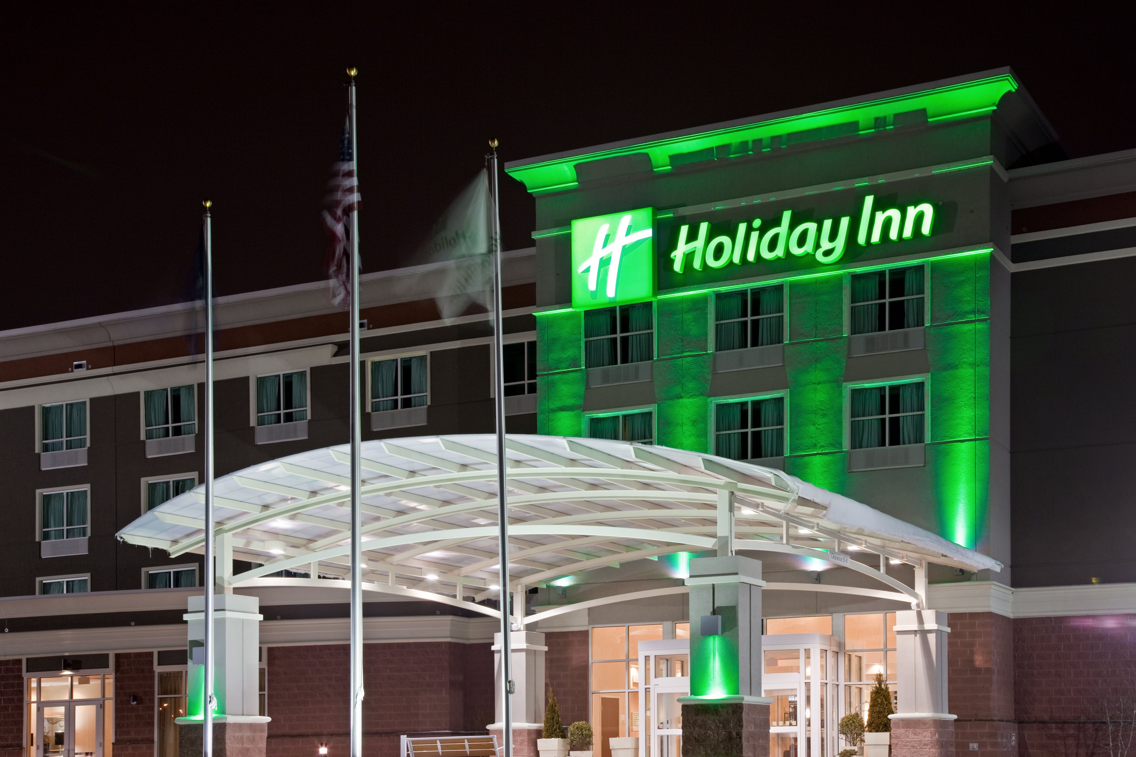 Un hotel Holiday Inn con un llamativo letrero verde, una entrada cubierta y un asta con la bandera estadounidense.
