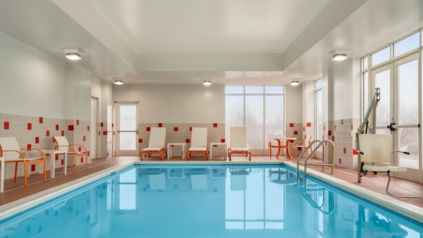 An indoor swimming pool with clear blue water, surrounded by white lounge chairs and a tiled wall with red accents.
