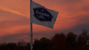 Eine amerikanische Flagge und ein Hampton Inn Schild an einem Fahnenmast vor einem Sonnenuntergangshimmel.