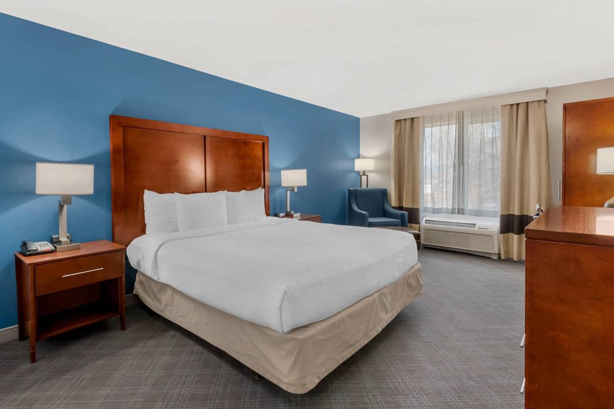 A hotel room with a large bed, two bedside tables with lamps, a chair, a desk, and a window with curtains.