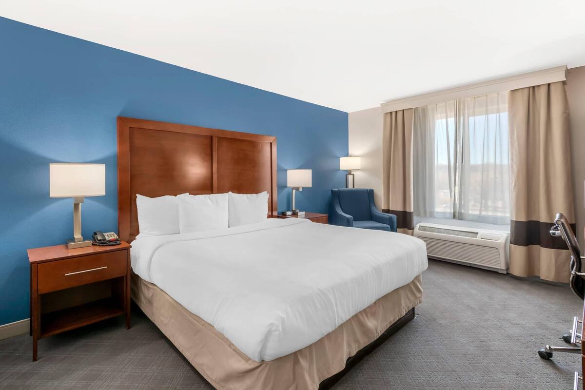 A hotel room with a large bed, two bedside tables with lamps, a chair, a window with curtains, and an air conditioning unit.