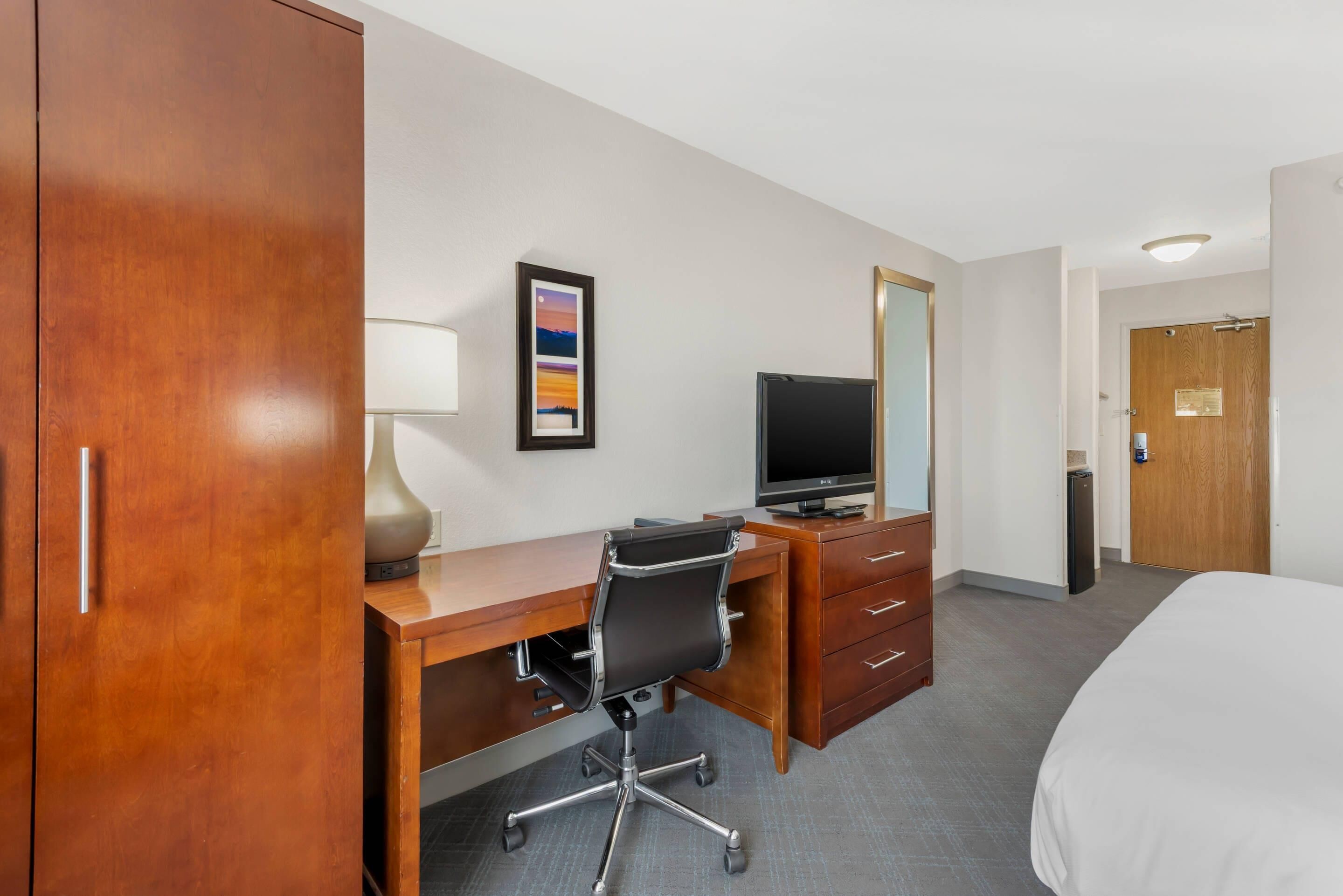 Quarto de hotel com uma escrivaninha de madeira, uma cadeira de escritório preta, uma televisão, uma cômoda e uma cama.