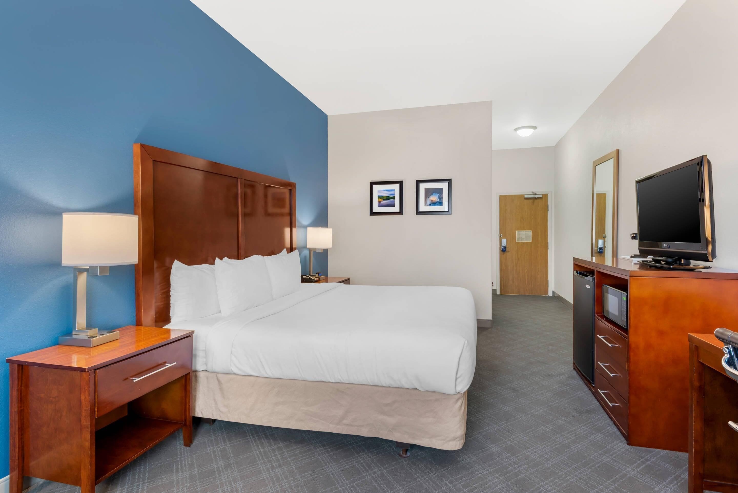 Una habitación de hotel con una cama grande, cabecera de madera, mesitas de noche con lámparas, cómoda con televisor y una puerta que da a otra habitación.