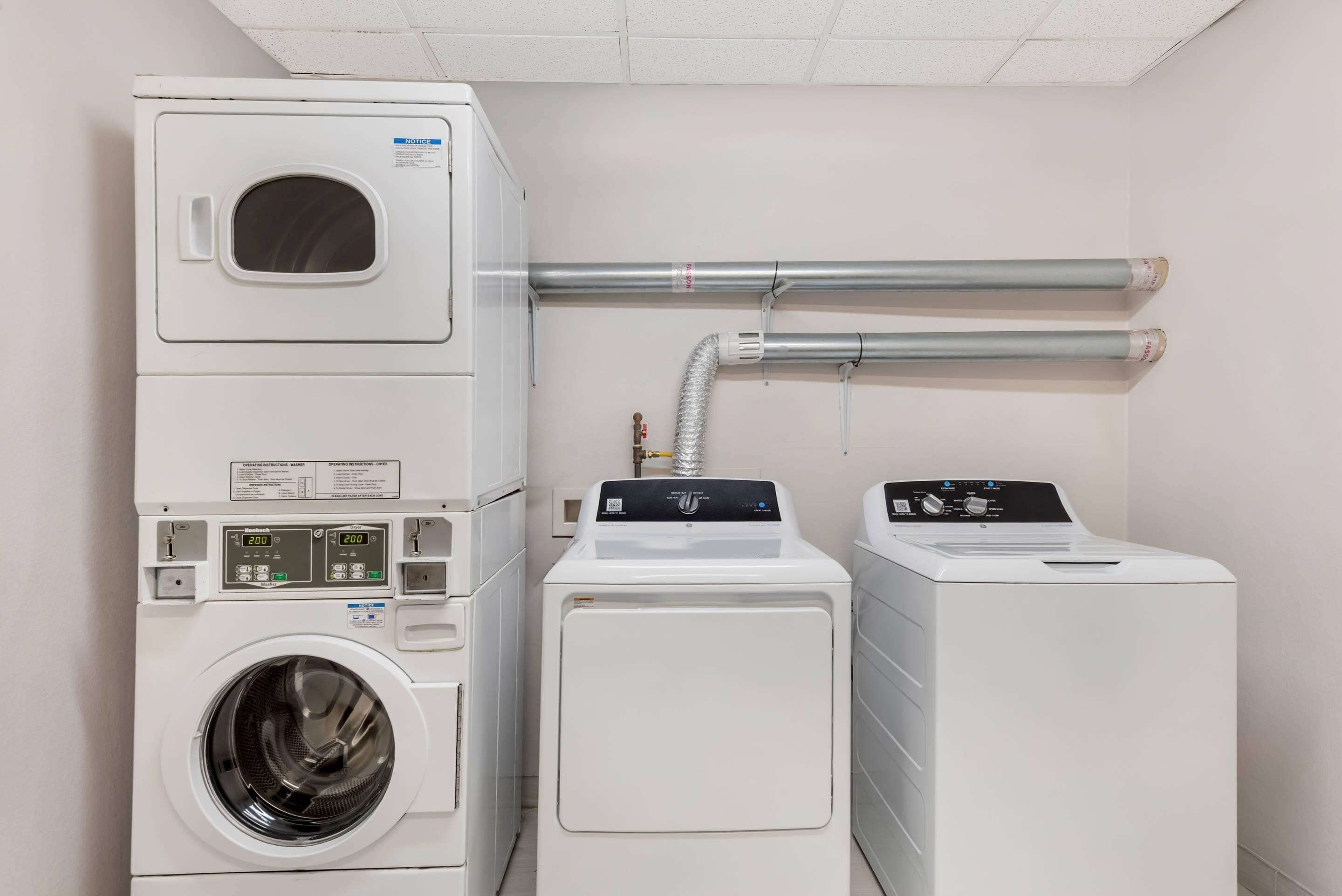 Un cuarto de lavandería con lavadoras y secadoras apiladas, conductos de ventilación y un panel de control.