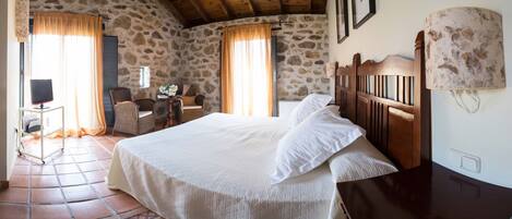 Un dormitorio con pared de piedra, una cama con ropa de cama blanca, cabecera de madera, un escritorio con una lámpara y una silla.