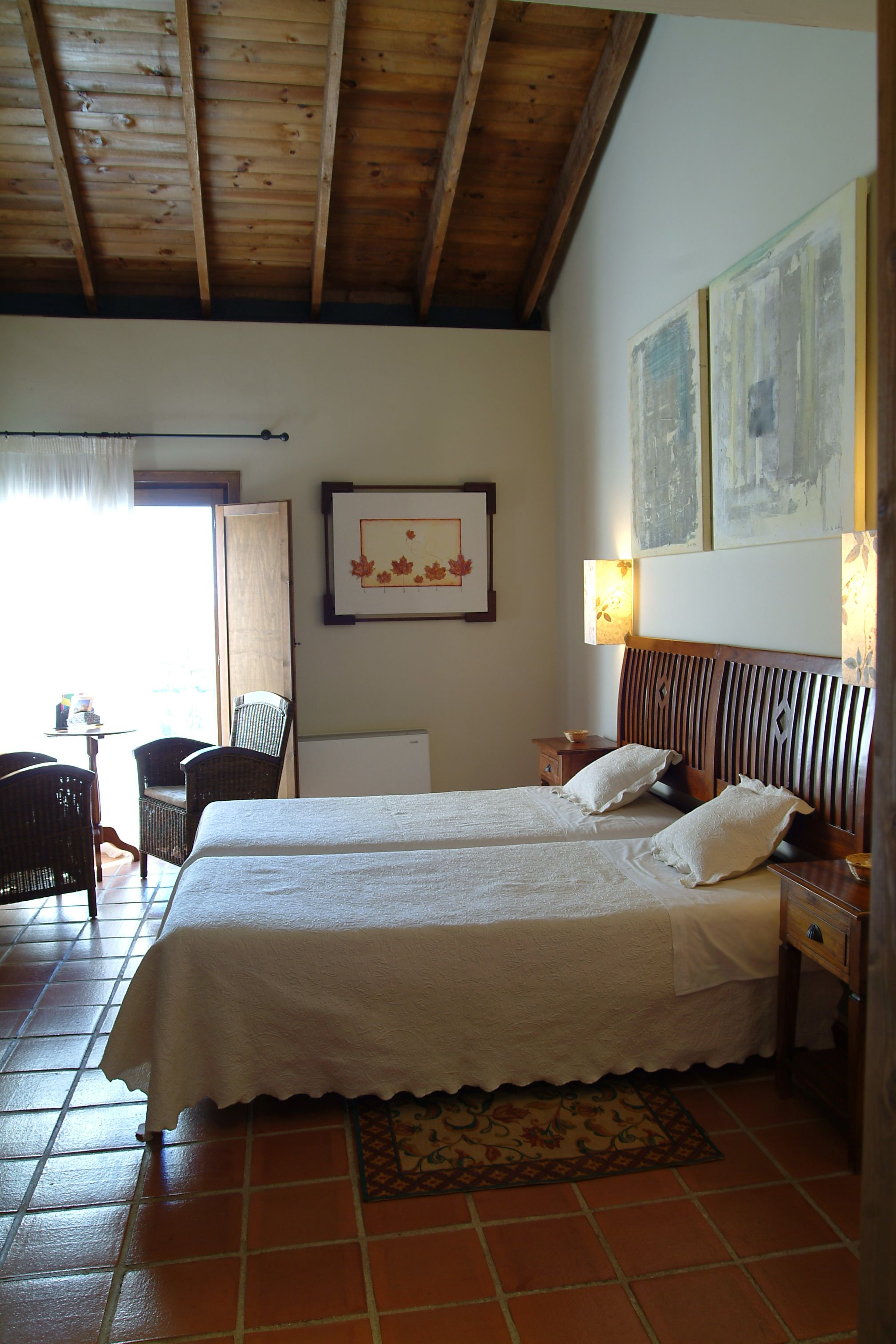 Un dormitorio con dos camas, techo de madera, suelo de baldosas, una pequeña mesa con sillas y un cuadro en la pared.