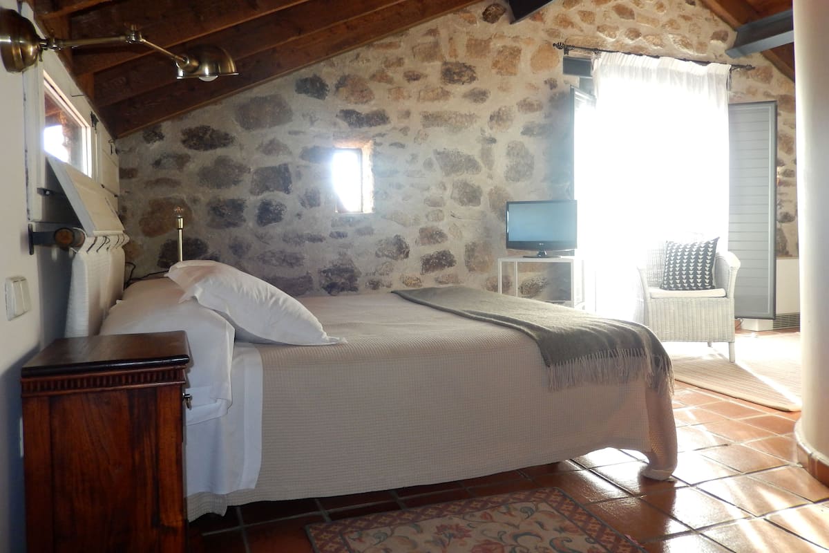 Un dormitorio con pared de piedra, una cama con ropa de cama blanca, un tocador de madera, un televisor sobre un mueble bajo y una silla de mimbre.