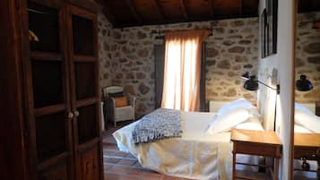 Una habitación rústica con pared de piedra, cama con cabecera blanca, armario de madera y una silla con cojín.