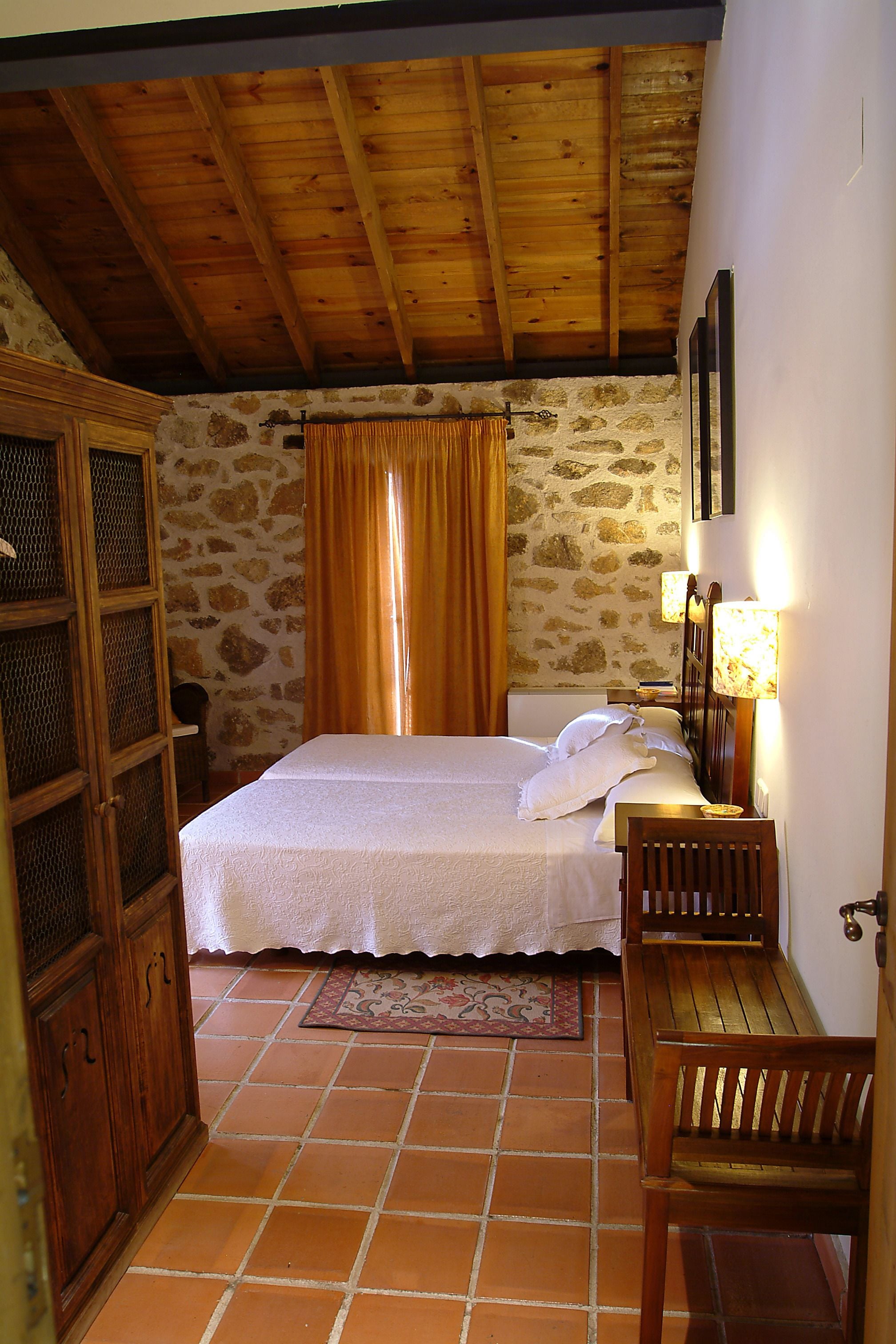 Un dormitorio con techo de madera, paredes de piedra, cama con ropa de cama blanca, mesita de noche con lámpara y una silla de madera.