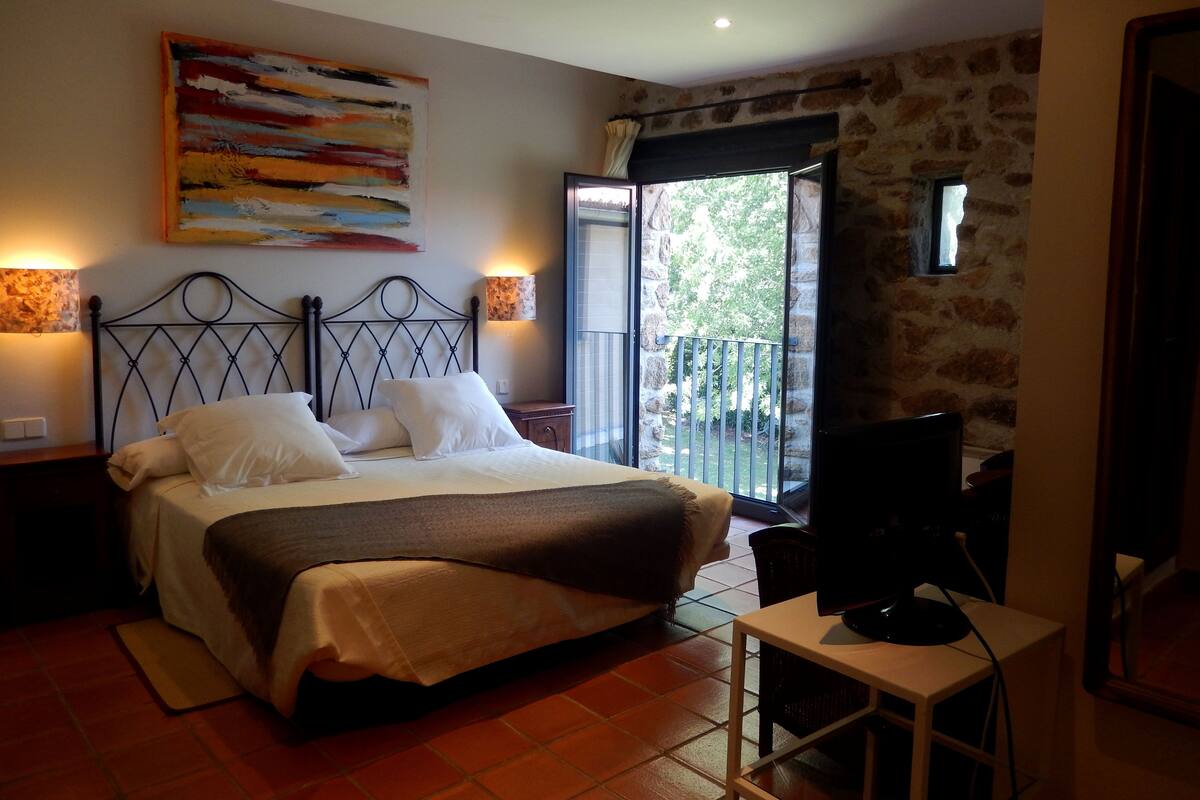 Un dormitorio con cama, escritorio, televisor, un cuadro y un balcón con vistas a árboles.