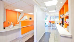 A modern reception area with a bright orange color scheme, a white reception desk, and a view of the outdoor seating area. - Première Classe Valence Sud (Valence)
