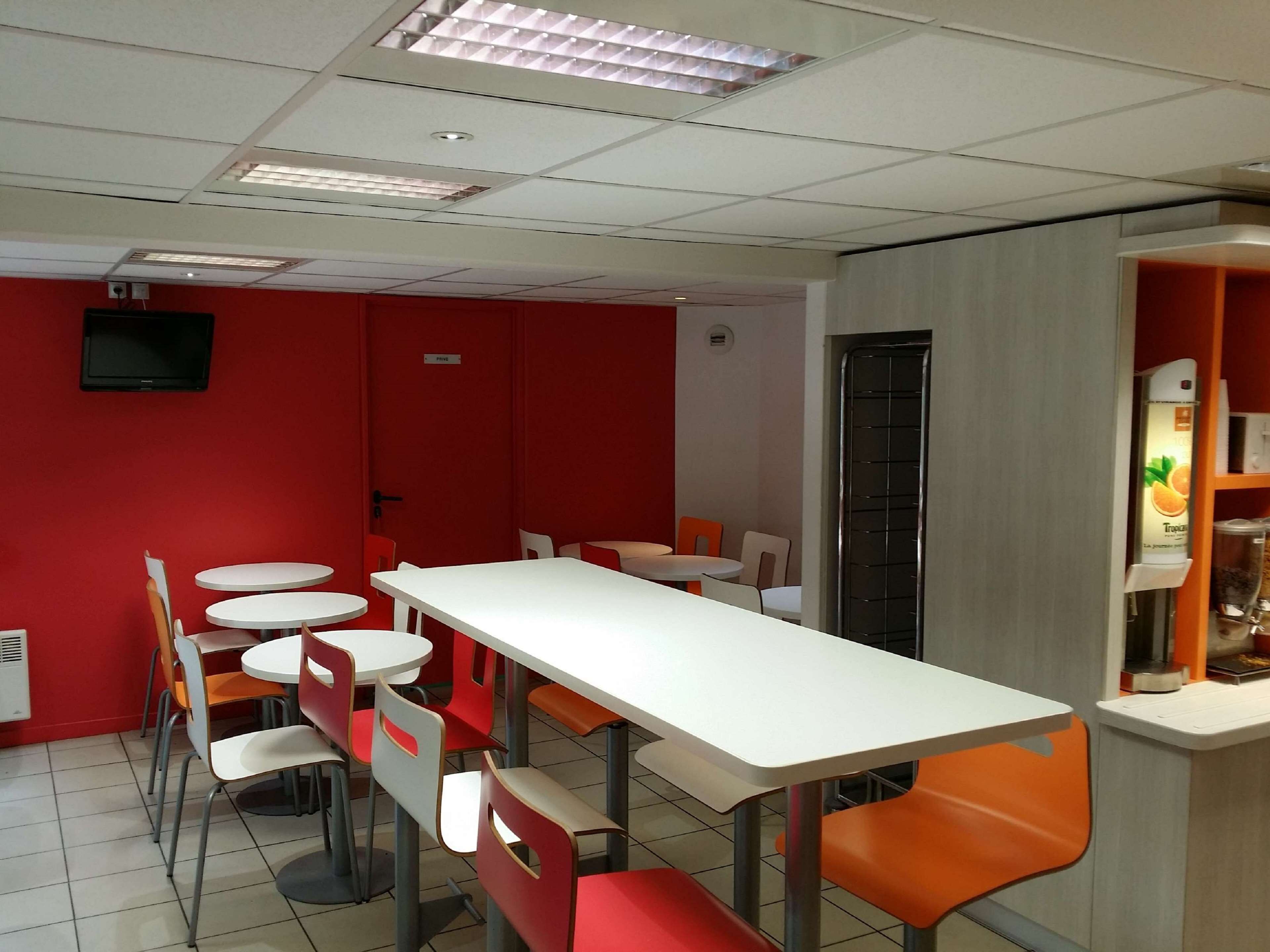 A modern dining area with white tables and orange chairs, red walls, and a vending machine.