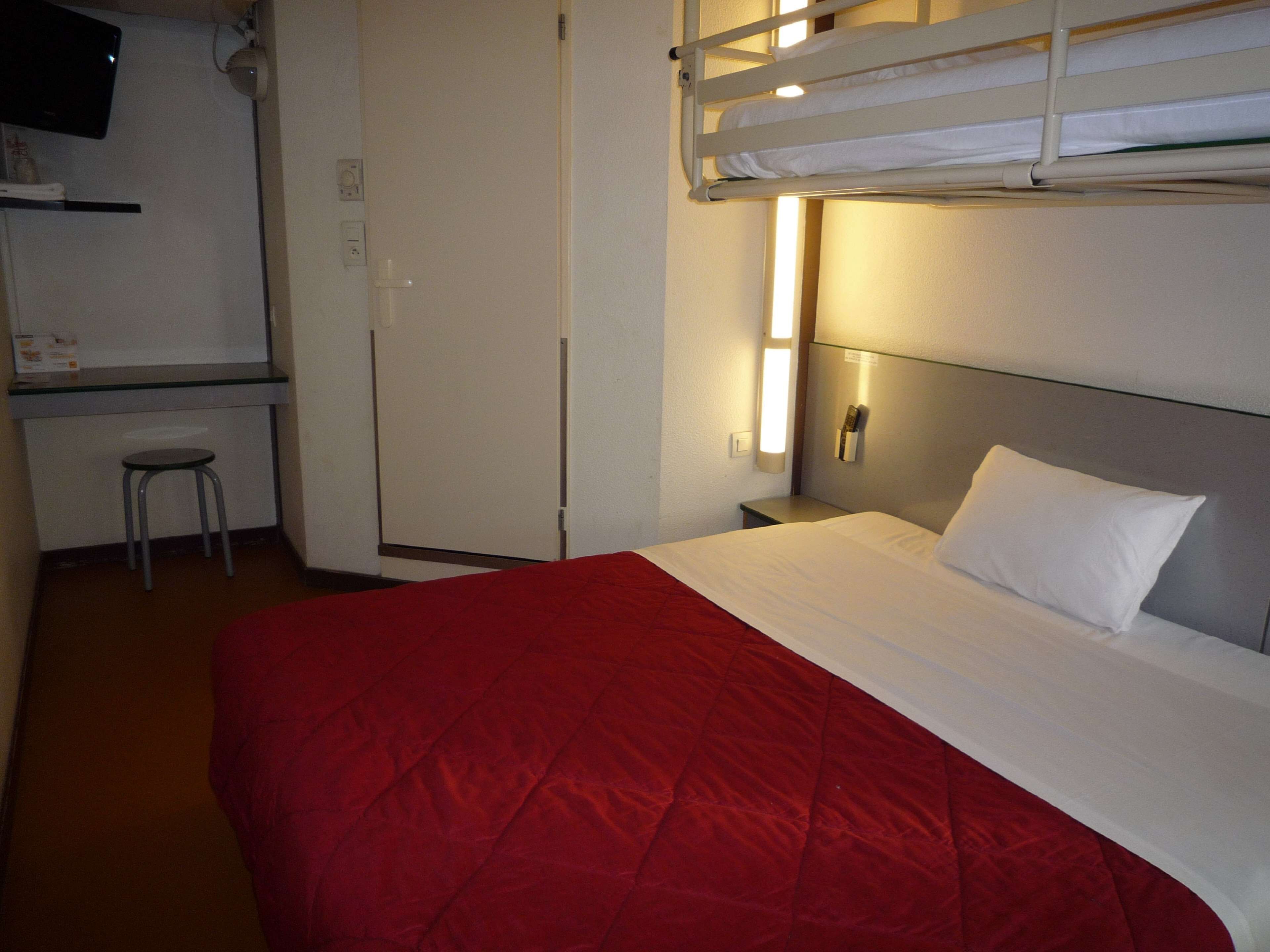 A hotel room with a bunk bed, a desk with a chair, a bedside table, and a TV mounted on the wall.
