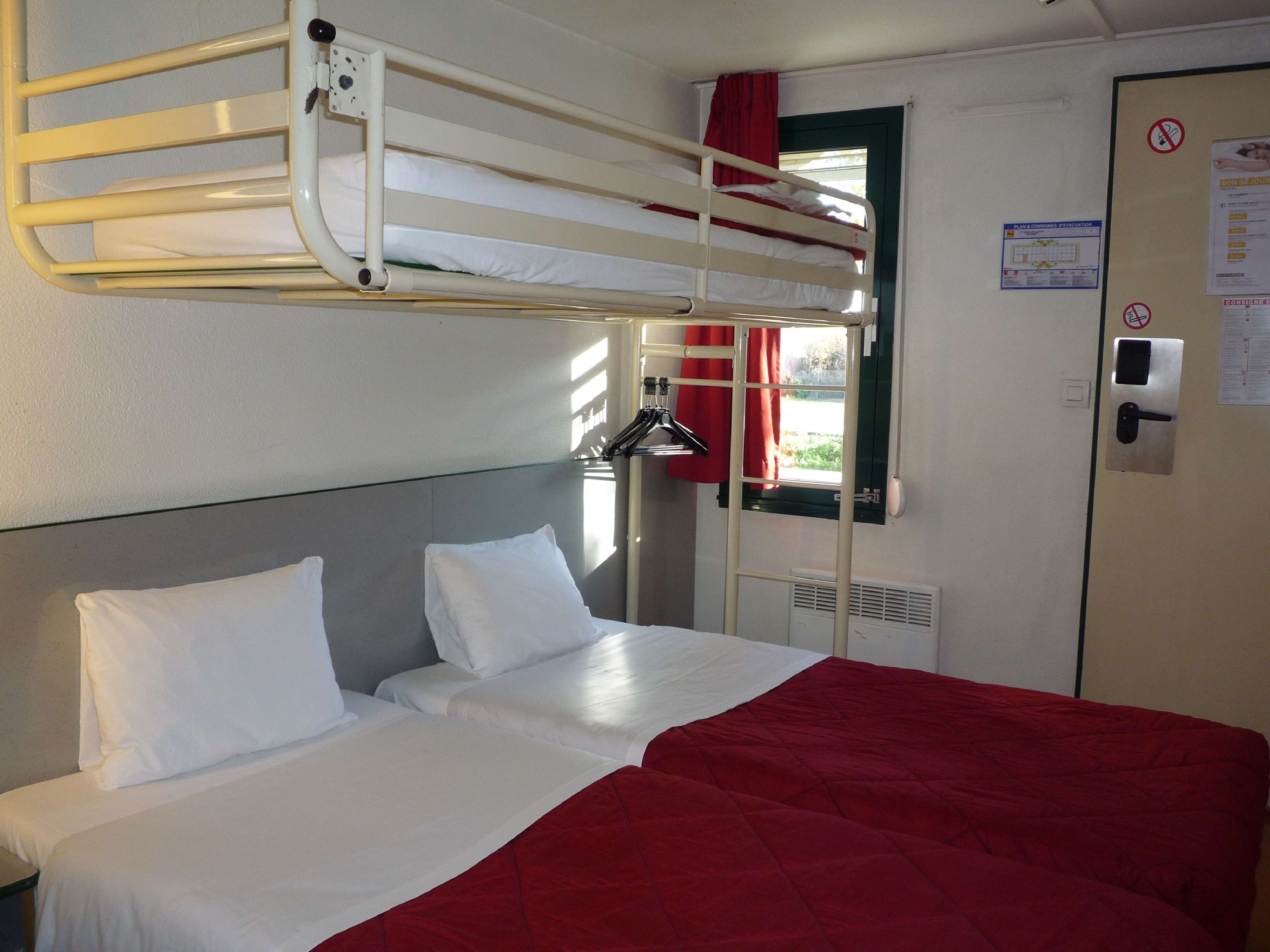 Una cama litera con una manta roja, una colcha blanca y una ventana con cortinas rojas.