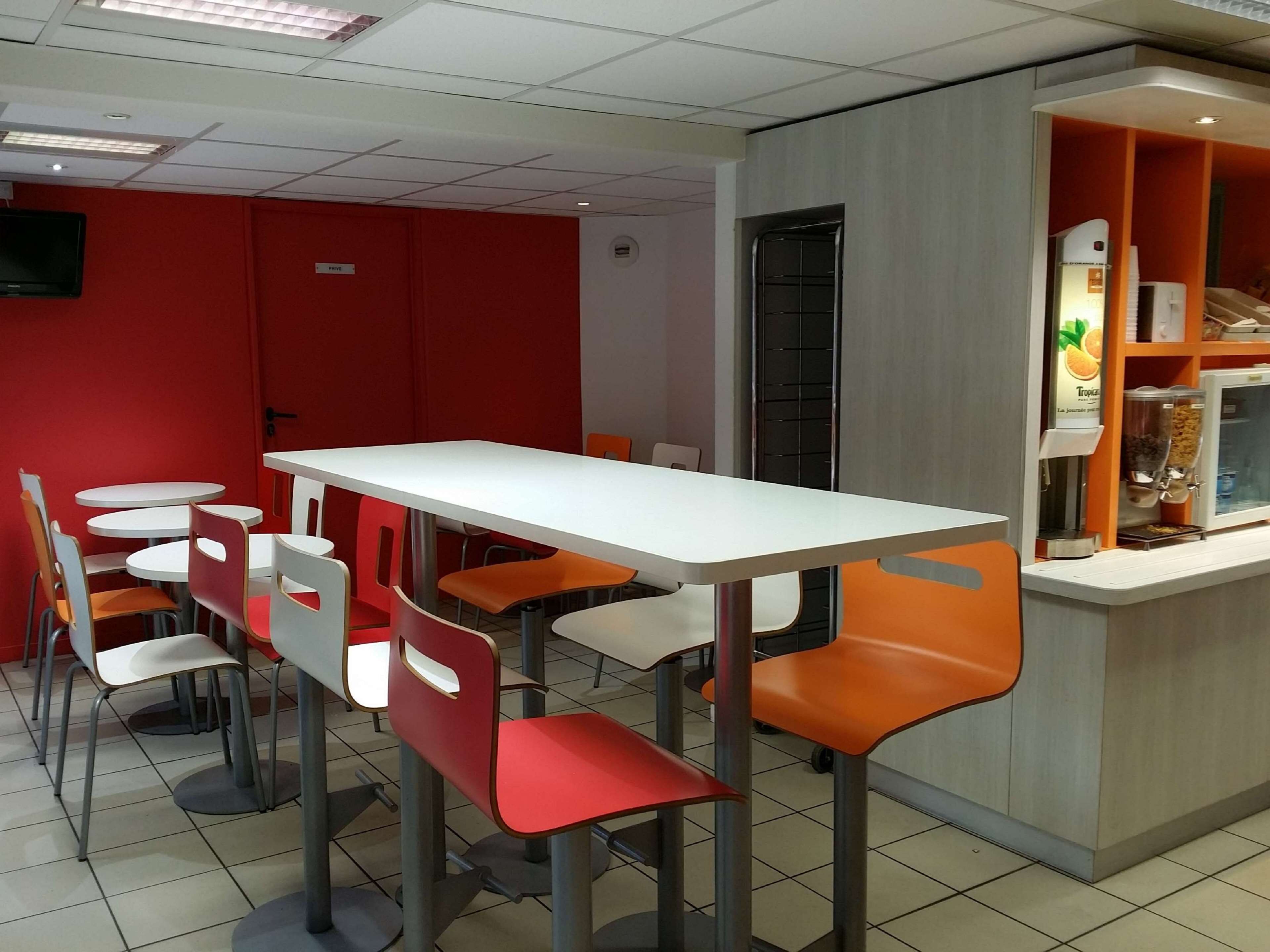 A modern dining area with a white table and orange chairs, a red wall, and a counter with a fruit dispenser.
