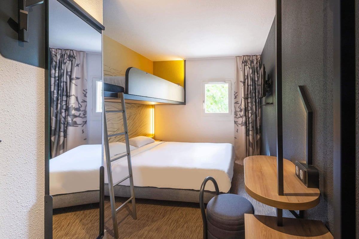 Habitación de hotel compacta con literas, un escritorio pequeño, una silla y una ventana con cortinas.